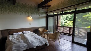 Ein Schlafzimmer mit einem großen Bett, einem kleinen runden Tisch, Korbstühlen und einem Balkon mit Blick auf Grünflächen.