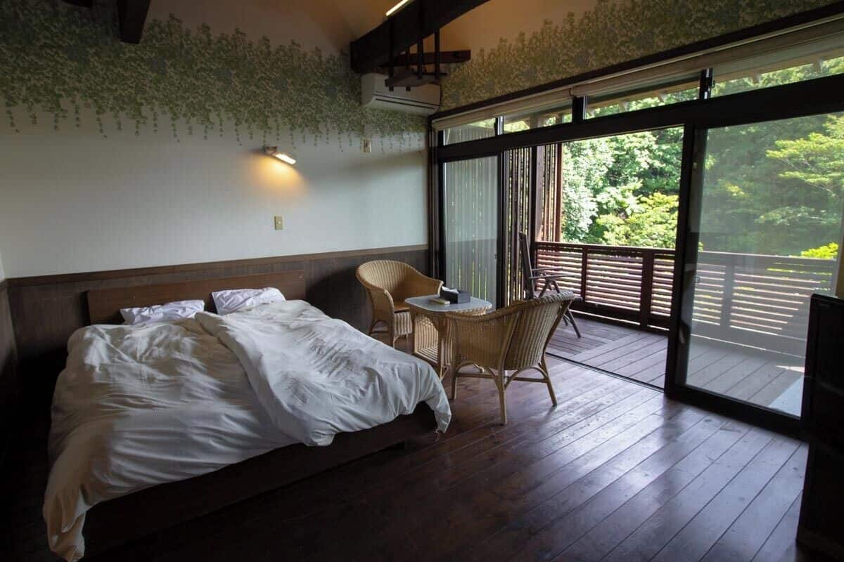 Un dormitorio con una cama grande, una mesita redonda pequeña, sillas de mimbre y un balcón con vistas a zonas verdes.
