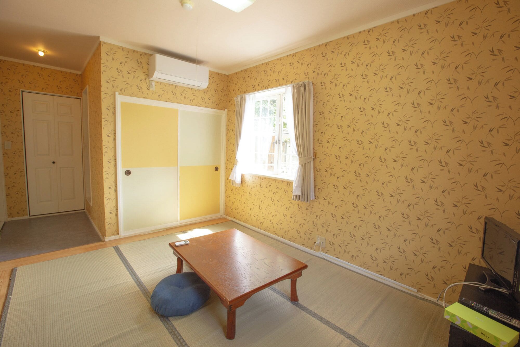 Une pièce avec un sol recouvert de tatamis, une table en bois, un coussin bleu, un téléviseur et une porte coulissante.