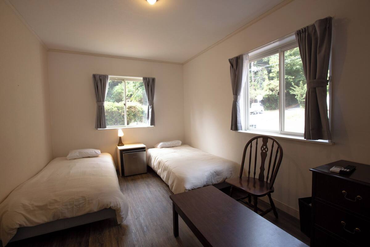 Una habitación pequeña y limpia con dos camas individuales, un escritorio, una silla y una ventana con cortinas.