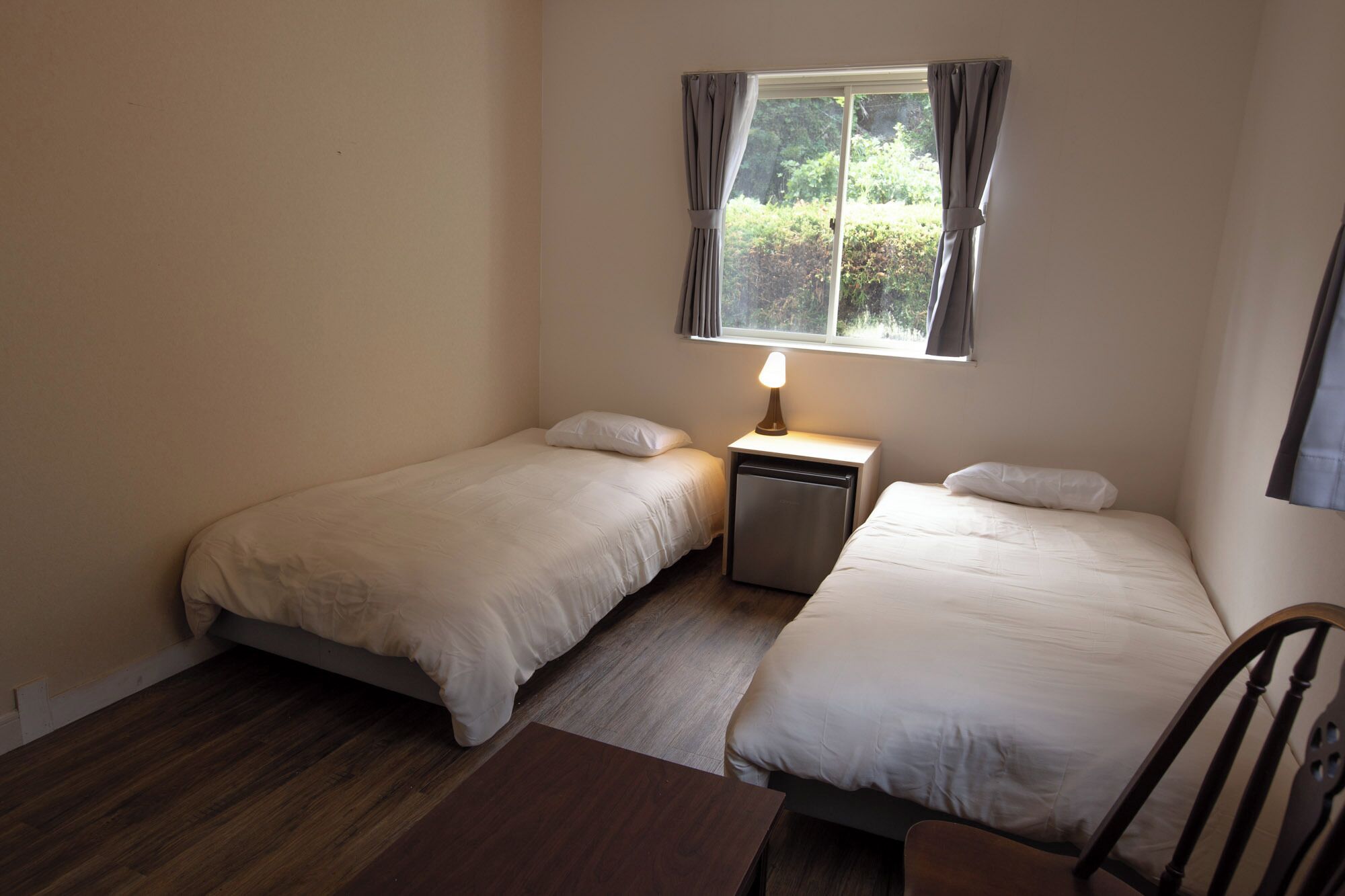 A small, clean room with two single beds, a nightstand with a lamp, a window with curtains, and a wooden floor.