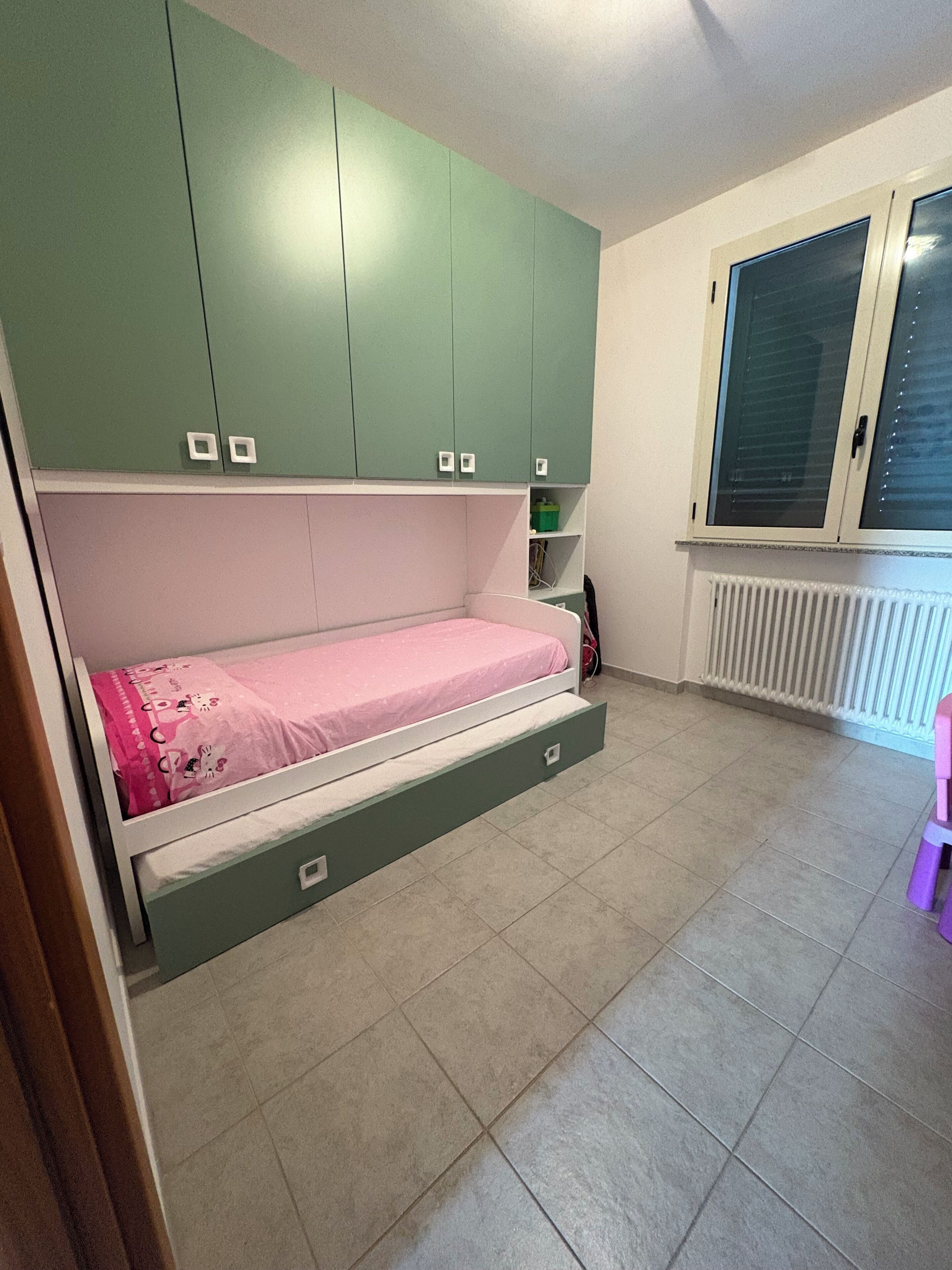 Un dormitorio con cama, armarios verdes, una ventana y un radiador.