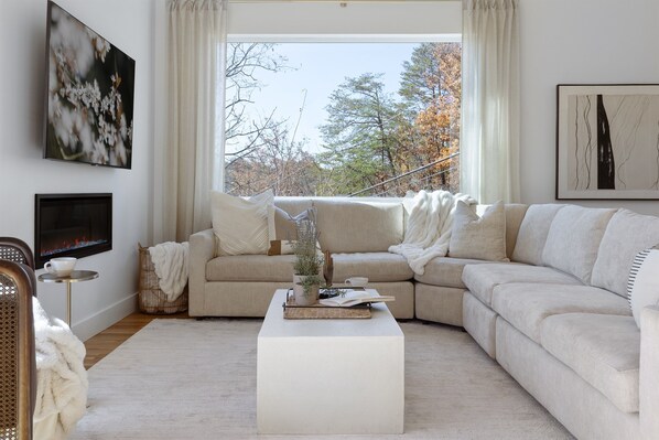 A modern living room with a beige sofa, a white coffee table, and a flat-screen TV mounted on the wall. - Luxe Asheville Retreats • Heated Pool + 2 Hot Tubs • Sleeps 16 • Views (Weaverville)
