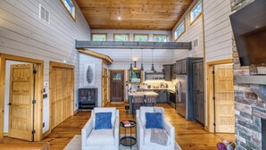A spacious living area with a high ceiling, wooden beams, a stone fireplace, and a kitchen with a central island. - Wellness Retreat! Peloton, Hot Tub, Sauna, Outdoor Gym, Views, and 2 King Suites (Cherry Log)