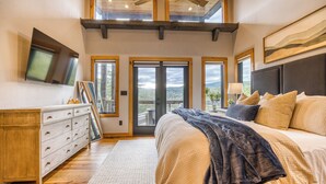 A bedroom with a large bed, a dresser, and a TV on the wall. There are multiple windows and a balcony. - Wellness Retreat! Peloton, Hot Tub, Sauna, Outdoor Gym, Views, and 2 King Suites (Cherry Log)