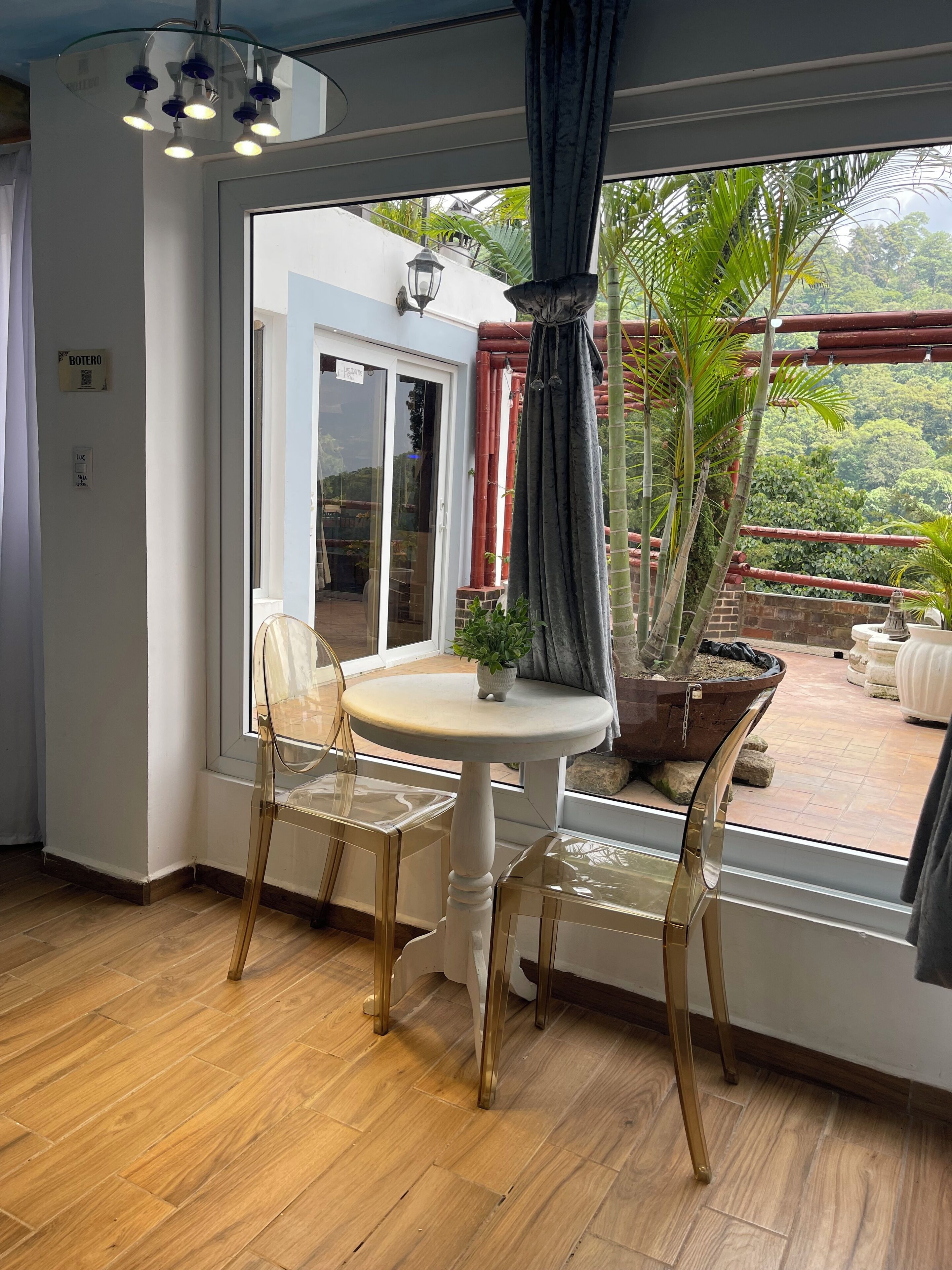 Ein Zimmer mit Holzboden, einem runden Tisch mit zwei Stühlen, einer Topfpflanze und Blick auf einen Balkon mit Pflanzen und einer roten Struktur.