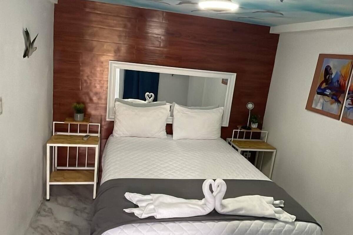 Une chambre d’hôtel comprenant un lit, une fresque représentant des oiseaux peinte au plafond, une tête de lit en bois, une table de chevet, une commode, un miroir et un tableau accroché au mur.