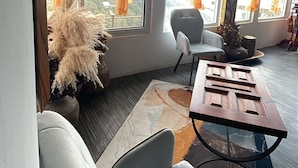 Une chambre moderne avec un canapé, une table et une vue sur les montagnes, visible à travers de grandes fenêtres ornées de rideaux orange.