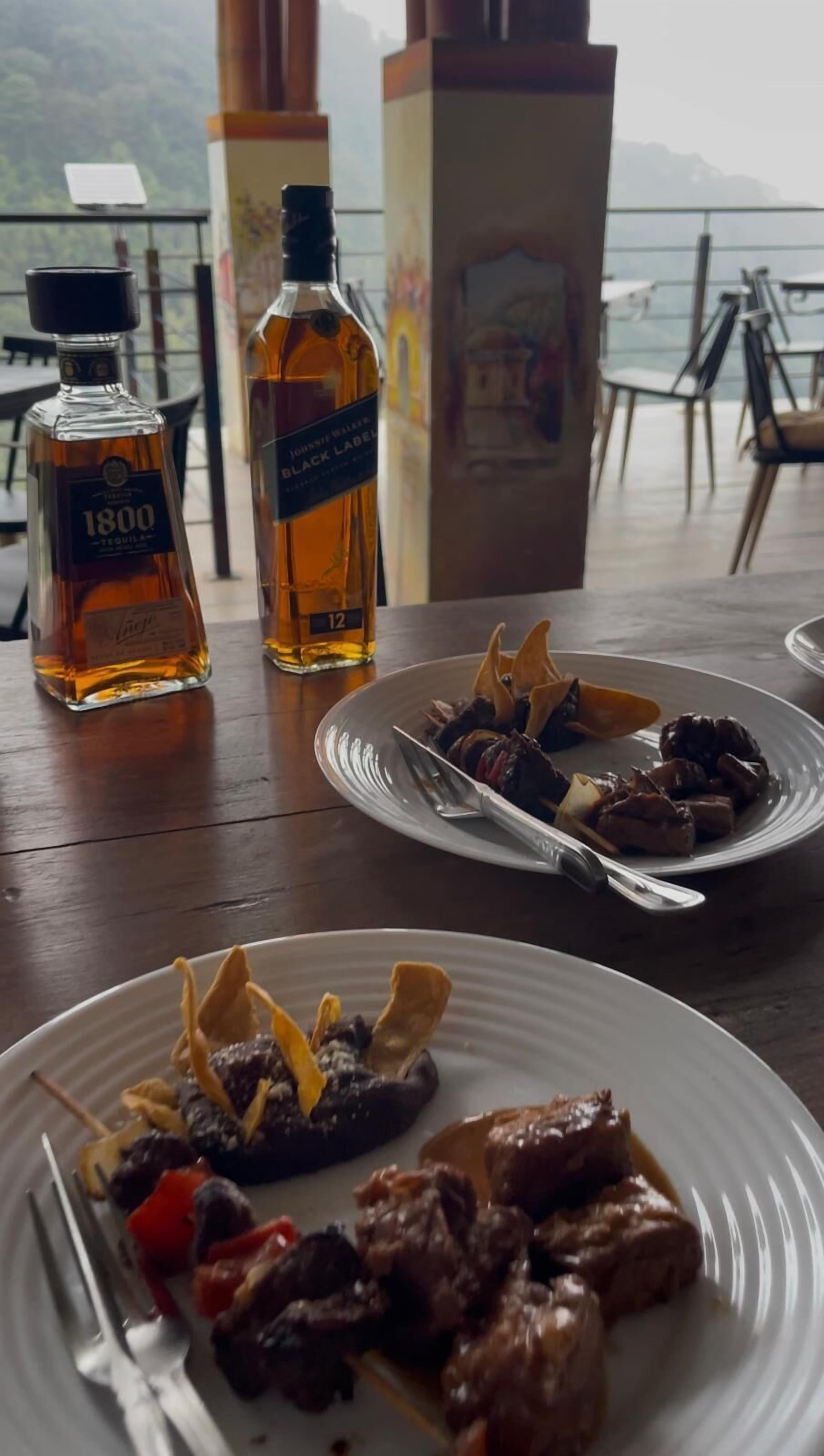 Une table avec deux assiettes de nourriture et deux bouteilles de spiritueux. L’endroit semble être un restaurant avec vue sur la mer.