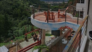 Une terrasse circulaire en plein air, dotée d’une rambarde en métal, entourée d’une végétation luxuriante et avec un paysage montagneux en arrière-plan.