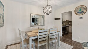 Een eethoek met een houten tafel en stoelen, een kroonluchter, een grote spiegel en een keuken op de achtergrond.