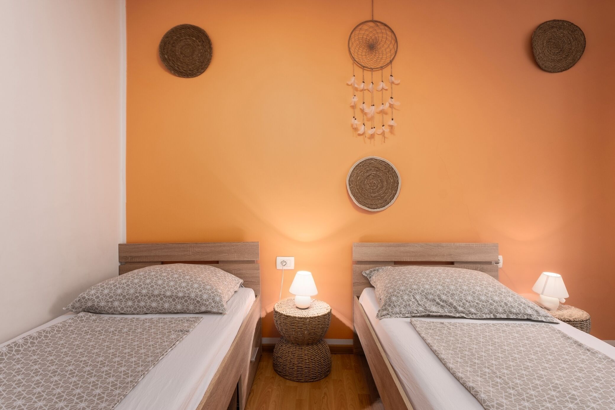 A room with two beds, each with a patterned bedspread and pillows, a bedside lamp, and a woven wall hanging.