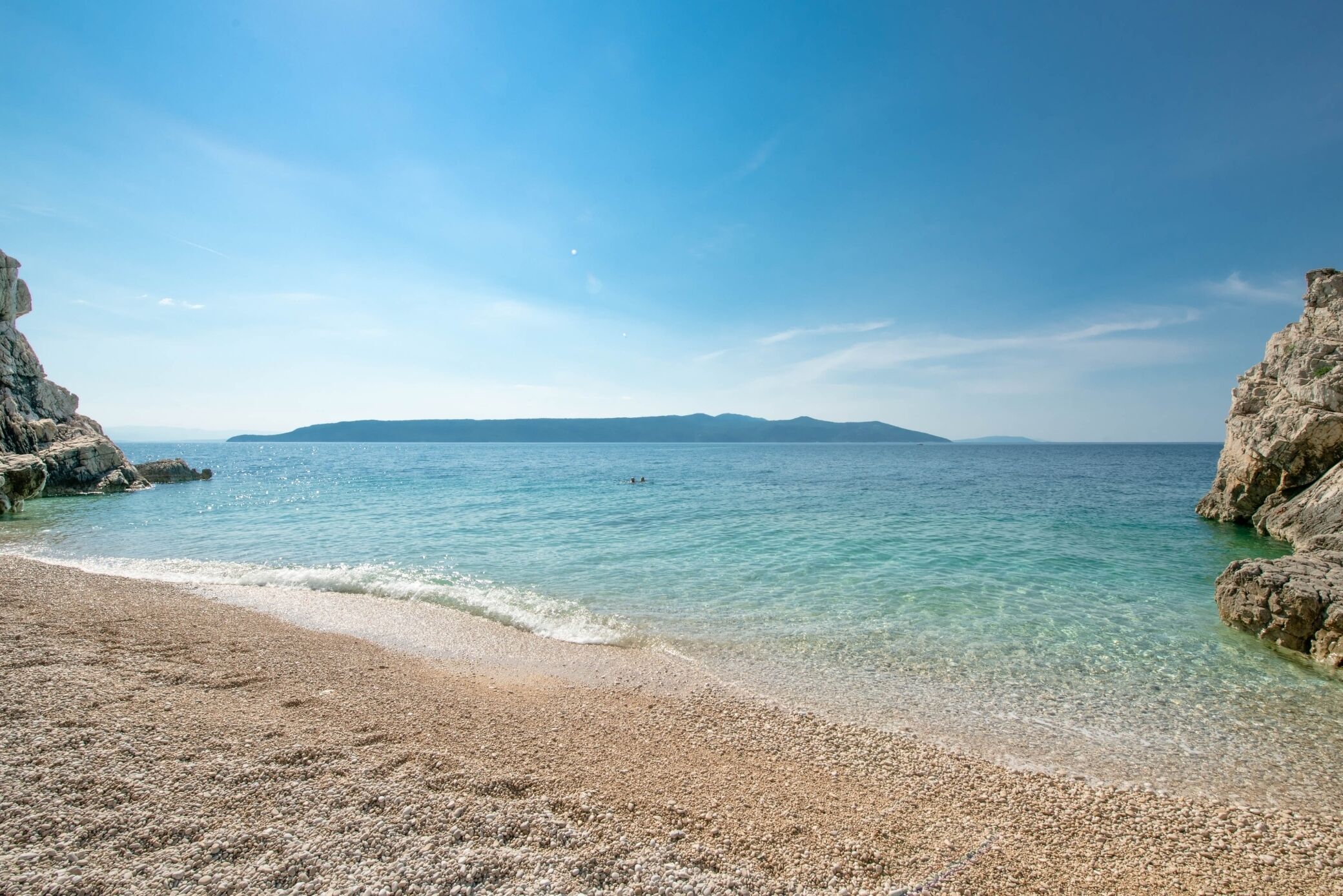A pebble beach with clear water and rocky cliffs.