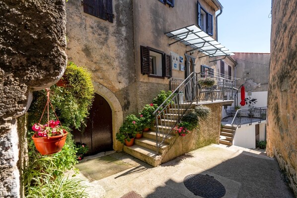 A narrow alleyway with a stone staircase leading to a second floor. There are potted plants along the steps and a hanging planter with flowers.