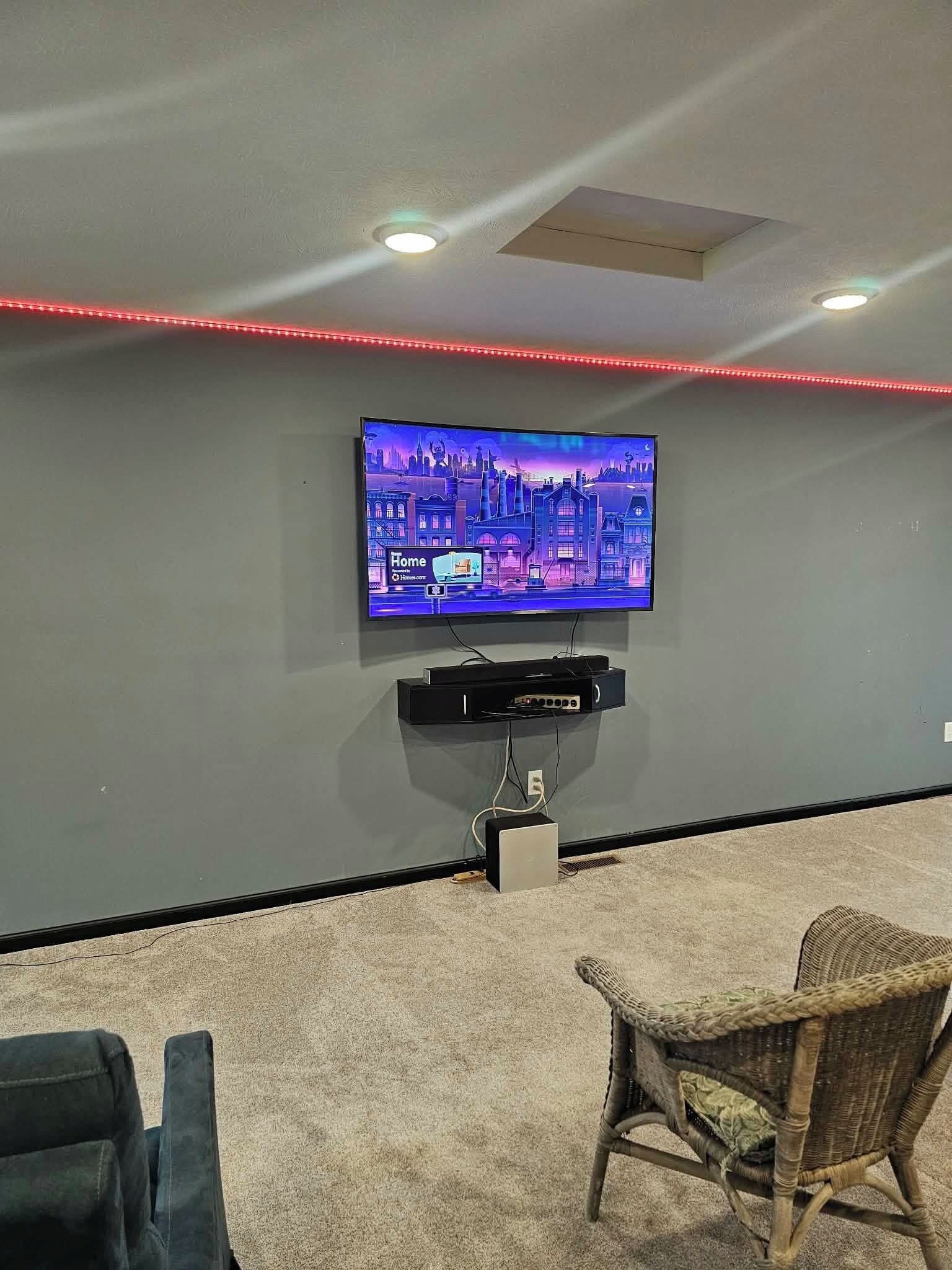 Sala de cinema com TV de tela plana na parede, projetor instalado no teto e fita de luz LED vermelha.