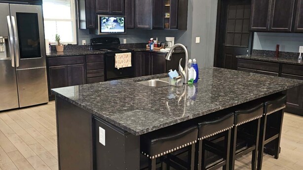 A modern kitchen with dark cabinetry, a granite island, stainless steel appliances, and a large window.