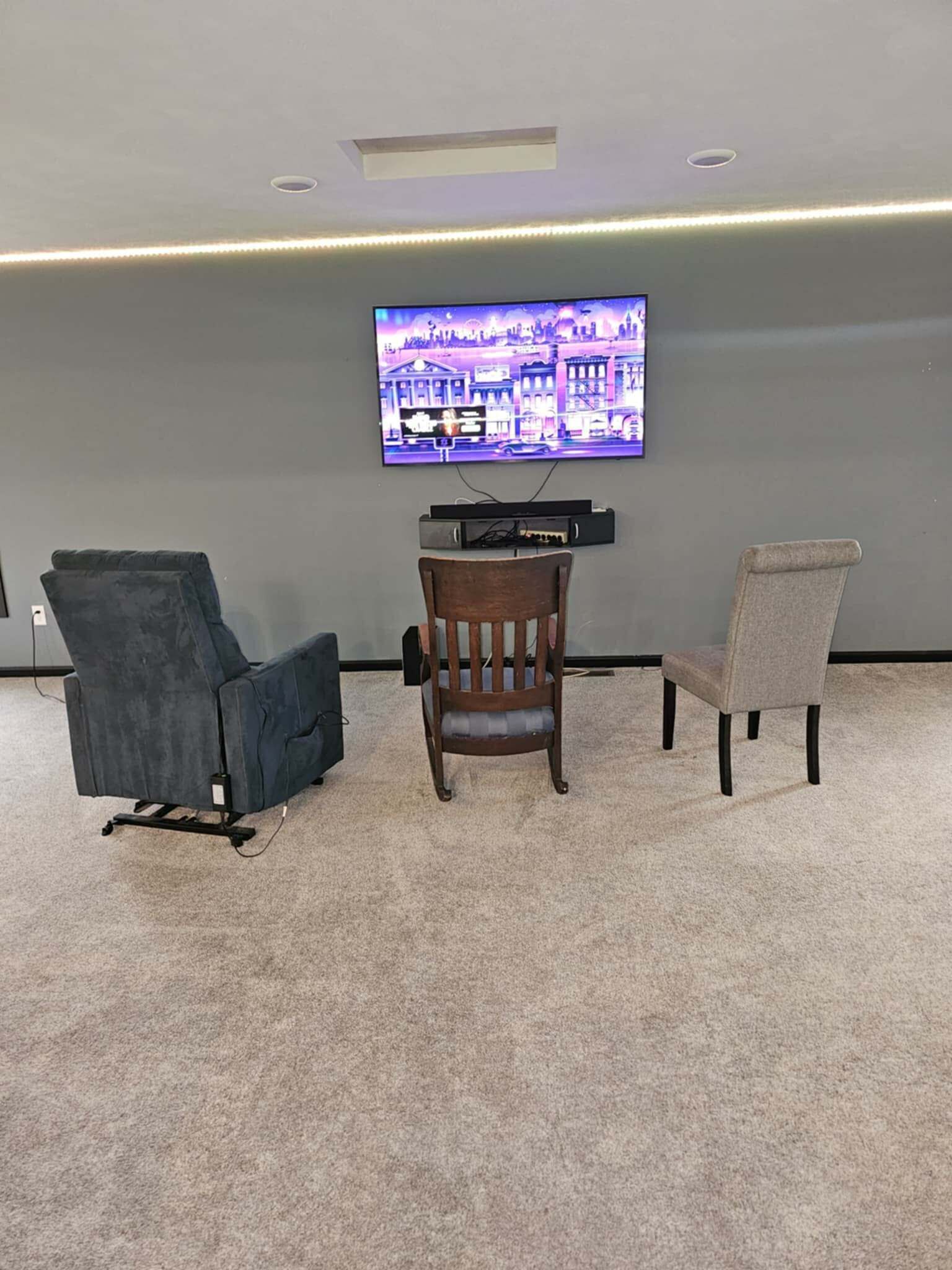 Una stanza con una TV a schermo piatto fissata al muro, una sedia di legno e un poltrona blu scuro.