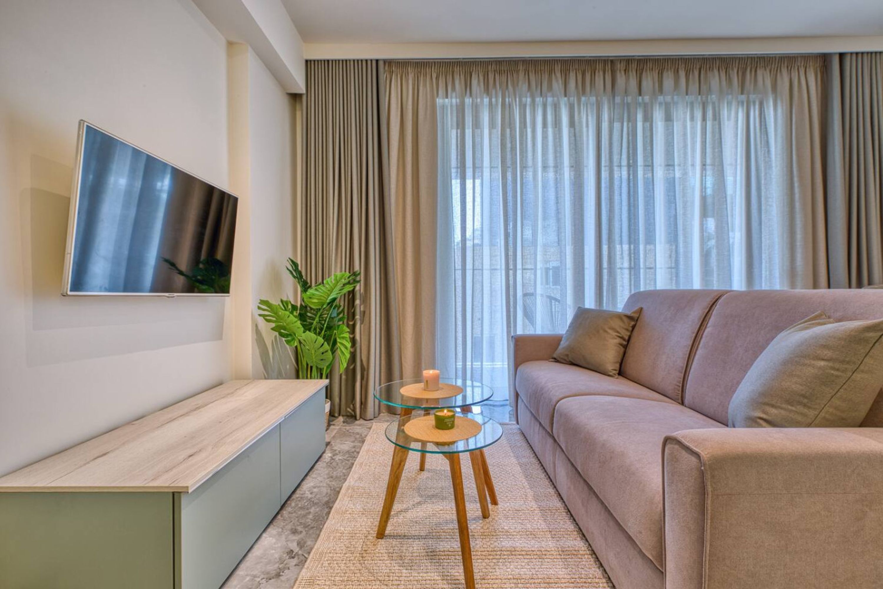 Una sala de estar moderna con un sofá, una mesa de centro de vidrio, un televisor montado en la pared y un amplio ventanal con cortinas.