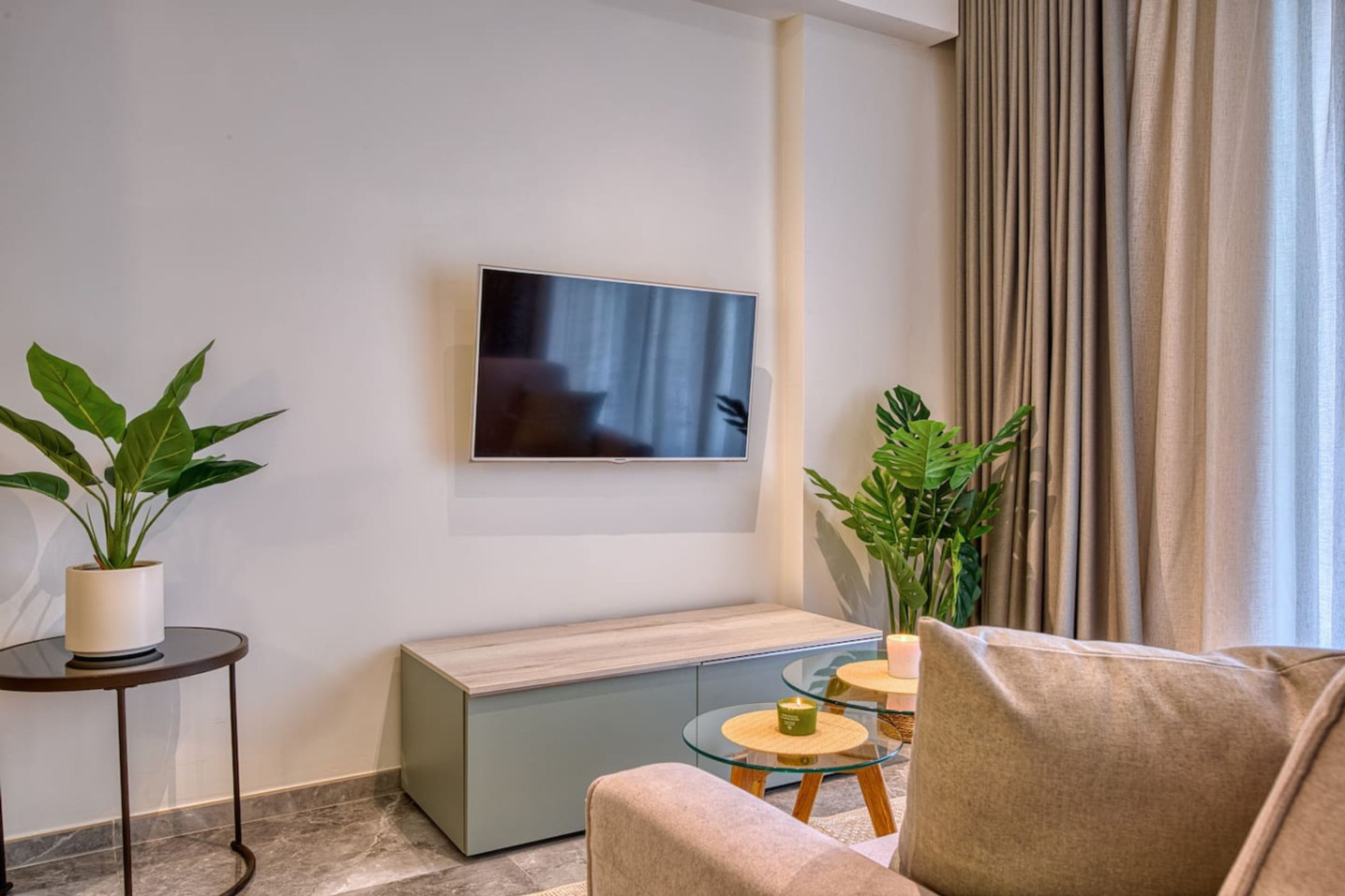 A modern living room with a flat-screen TV mounted on the wall, a sofa, a small coffee table, and two potted plants.