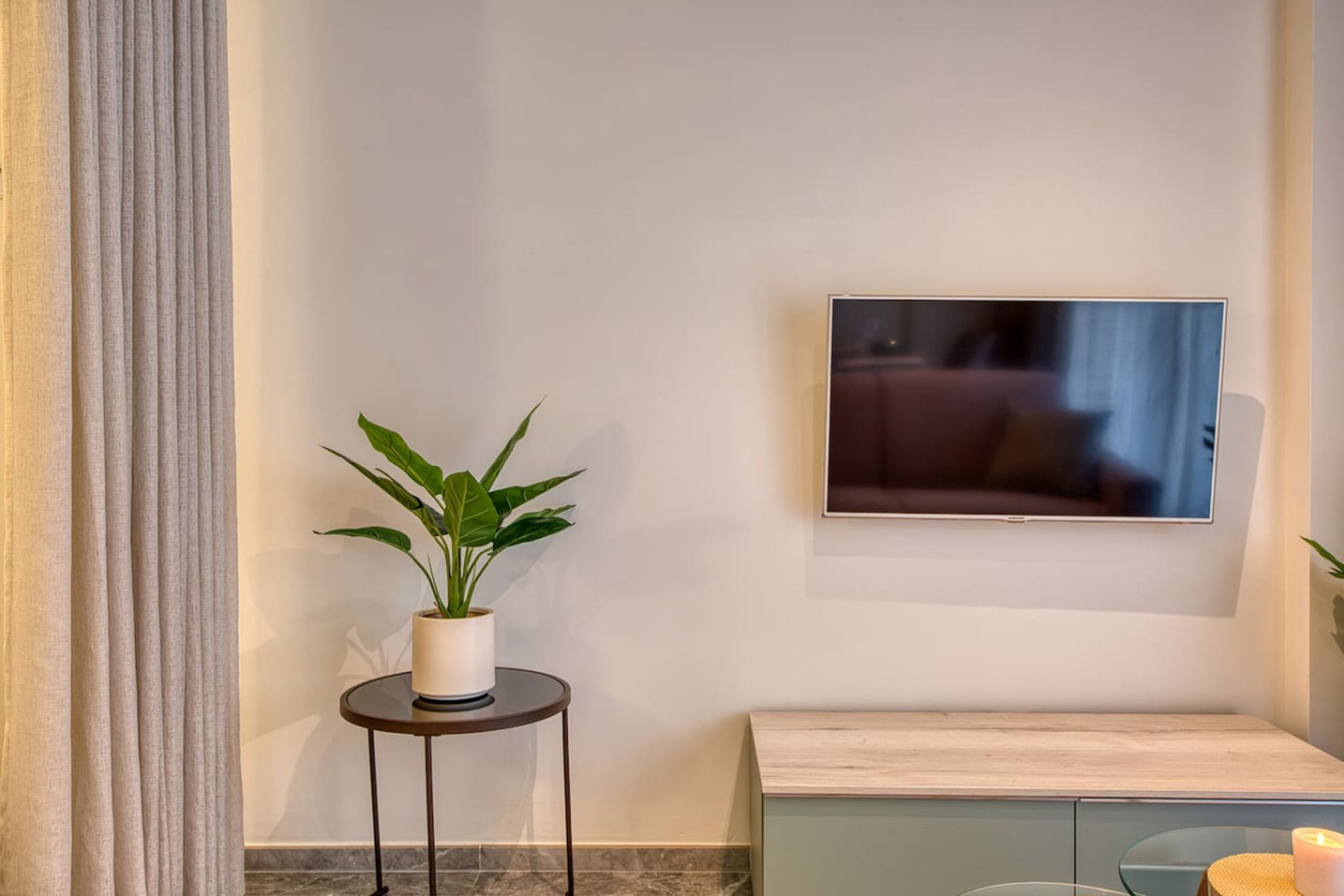 A room with a TV mounted on the wall, a small round table with a plant, and a light-colored cabinet.