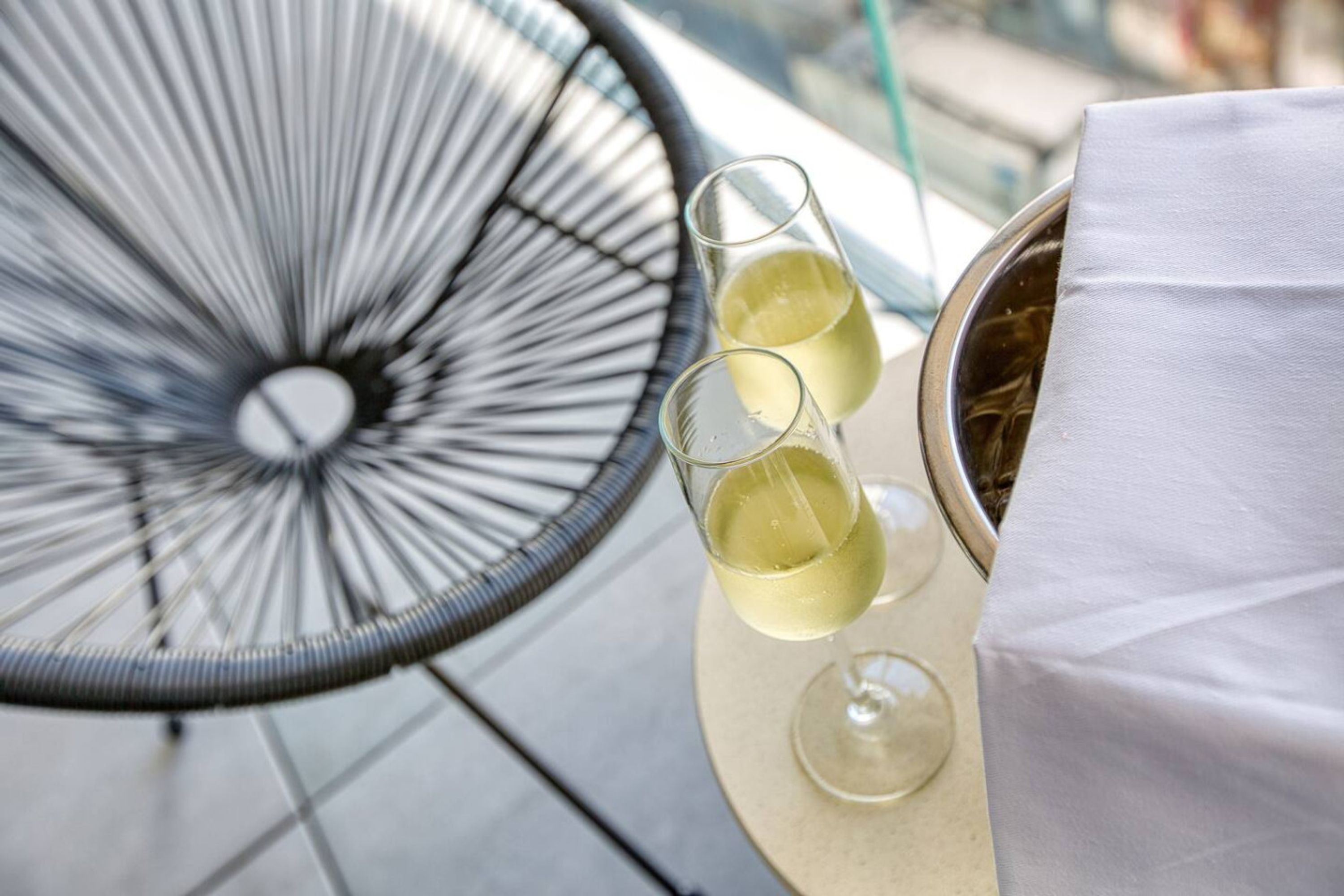 A round table with two glasses of white wine and a metal container, likely a wine cooler, on a balcony with a metal chair.