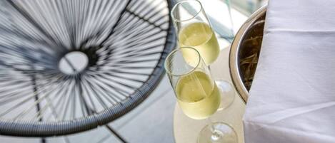 A round table with two glasses of white wine and a metal container, likely a wine cooler, on a balcony with a metal chair.