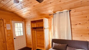 Interior de una cabaña de madera con ventilador de techo, un sofá, una ventana y una puerta.