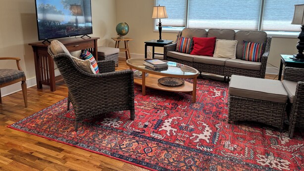 A living room with a television, wicker furniture, a red patterned rug, and a window with blinds.