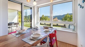 A dining area with a wooden table set for two, large windows with a view of greenery and buildings, and pendant lights hanging above. - Nice apartment for 2 guests with WIFI, TV and terrace (Ossiachberg)