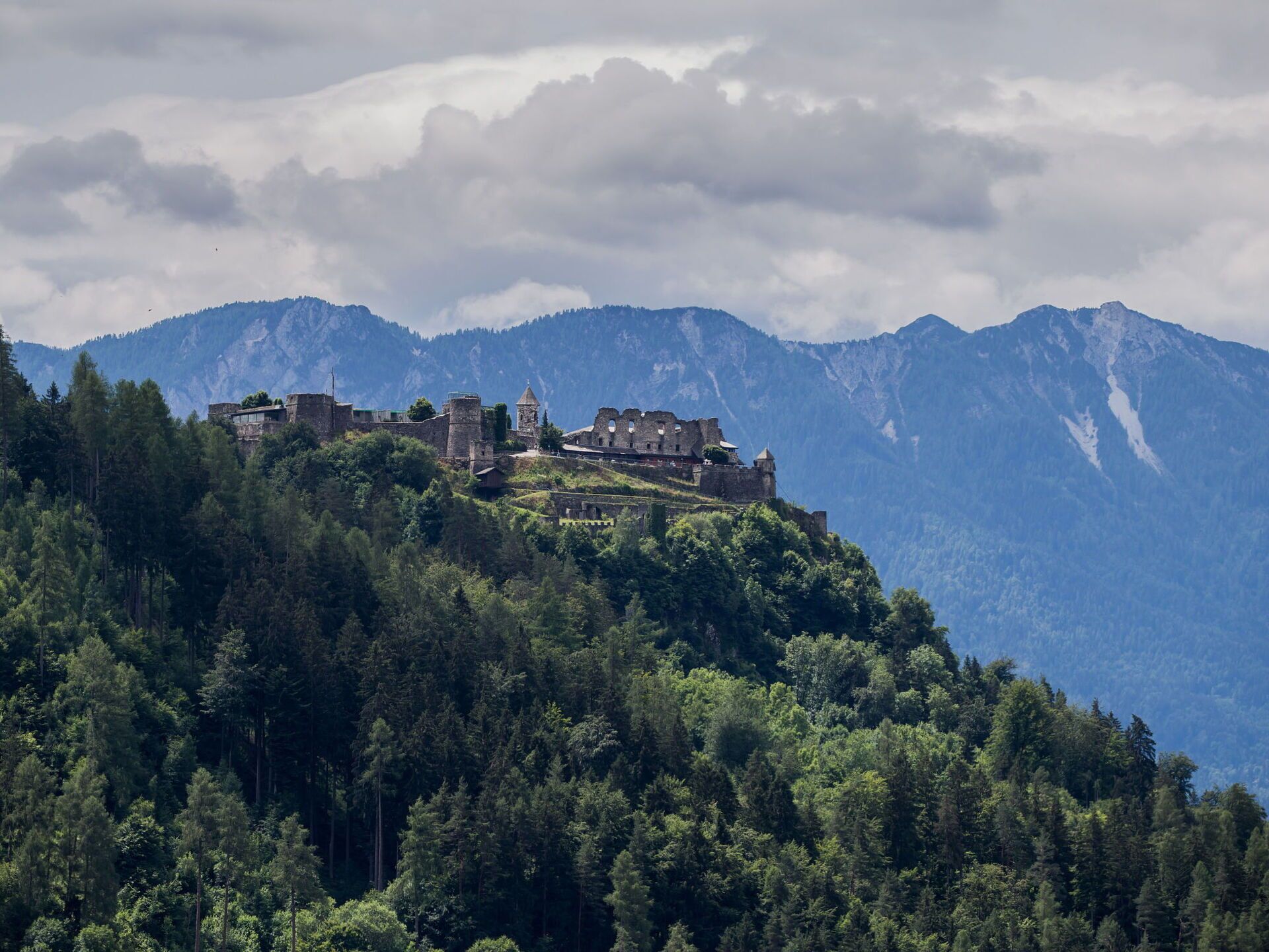 Un château historique perché sur une colline, entouré d’une forêt dense et avec des montagnes en arrière-plan.