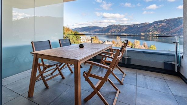 A balcony with a wooden table and chairs, offering a view of a lake and mountains. - Nice apartment for 2 guests with WIFI, TV and terrace (Ossiachberg)