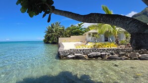 A beachfront property with clear water, a stone wall, and a small building with a thatched roof. - Villa Kahia by Tahiti Homes (Windward Islands)