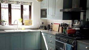 A kitchen with teal cabinets, a stove, a sink, and a window with curtains. - Atworth Haven: 4-Bed Home with Garden (Atworth)
