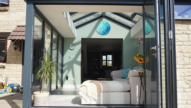 A modern glass-enclosed living area with a sofa, a coffee table, and a potted plant. - Atworth Haven: 4-Bed Home with Garden (Atworth)