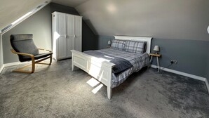 A bedroom with a sloped ceiling, a bed with a plaid bedspread, a rocking chair, and a wardrobe. - Atworth Haven: 4-Bed Home with Garden (Atworth)