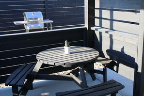 Un patio de madera con una mesa redonda y bancos, una parrilla y un cactus.