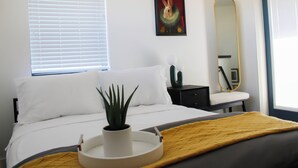 A modern bedroom with a bed, a bedside table, a potted plant, a painting, and a mirror.