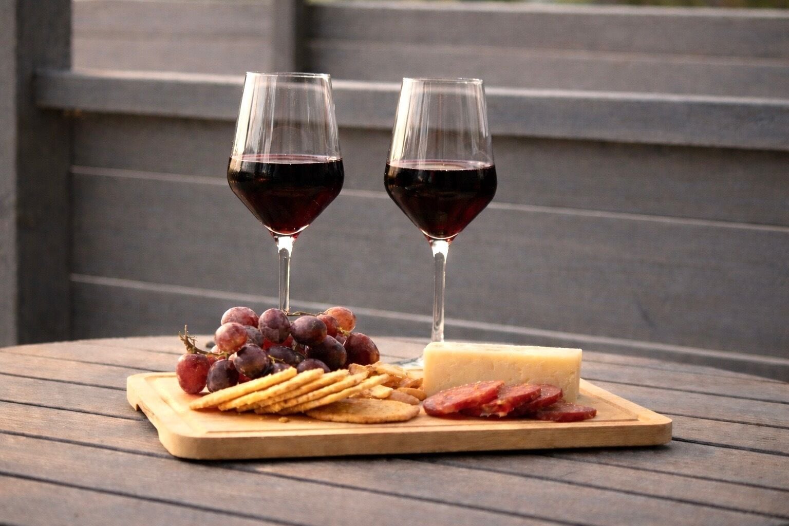 Et træbord med et ostebræt, druer, kiks og skivet salamipølse. To glas rødvin er også synlige.