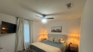 Een slaapkamer met een bed, twee bedlampen, een plafondventilator, een bevestigde televisie en een raam met gordijnen.