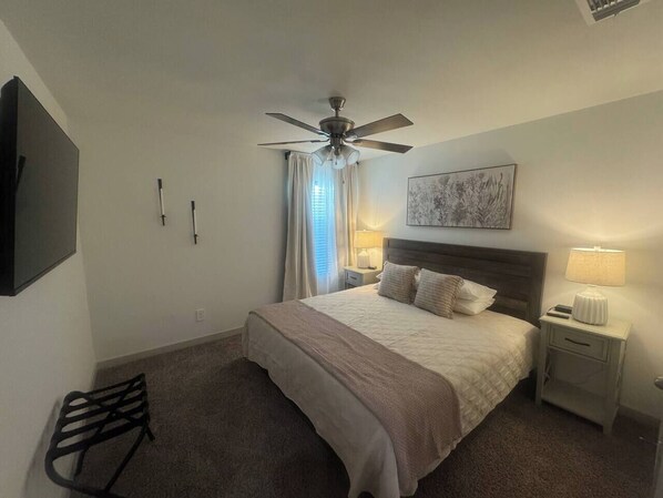 A hotel room with a bed, a ceiling fan, a nightstand with a lamp, a chair, and a painting on the wall. - Fifty5 Modern Westmont Retreat (Lubbock)