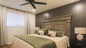Ein ordentlich eingerichtetes Schlafzimmer mit einem Bett, zwei Nachttischen, einem Deckenventilator und einem Fenster mit Vorhängen.