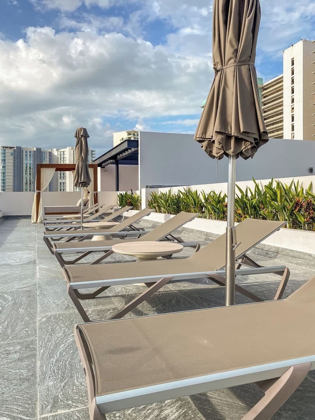 A rooftop terrace with lounge chairs and umbrellas, a pool, and a view of high-rise buildings.