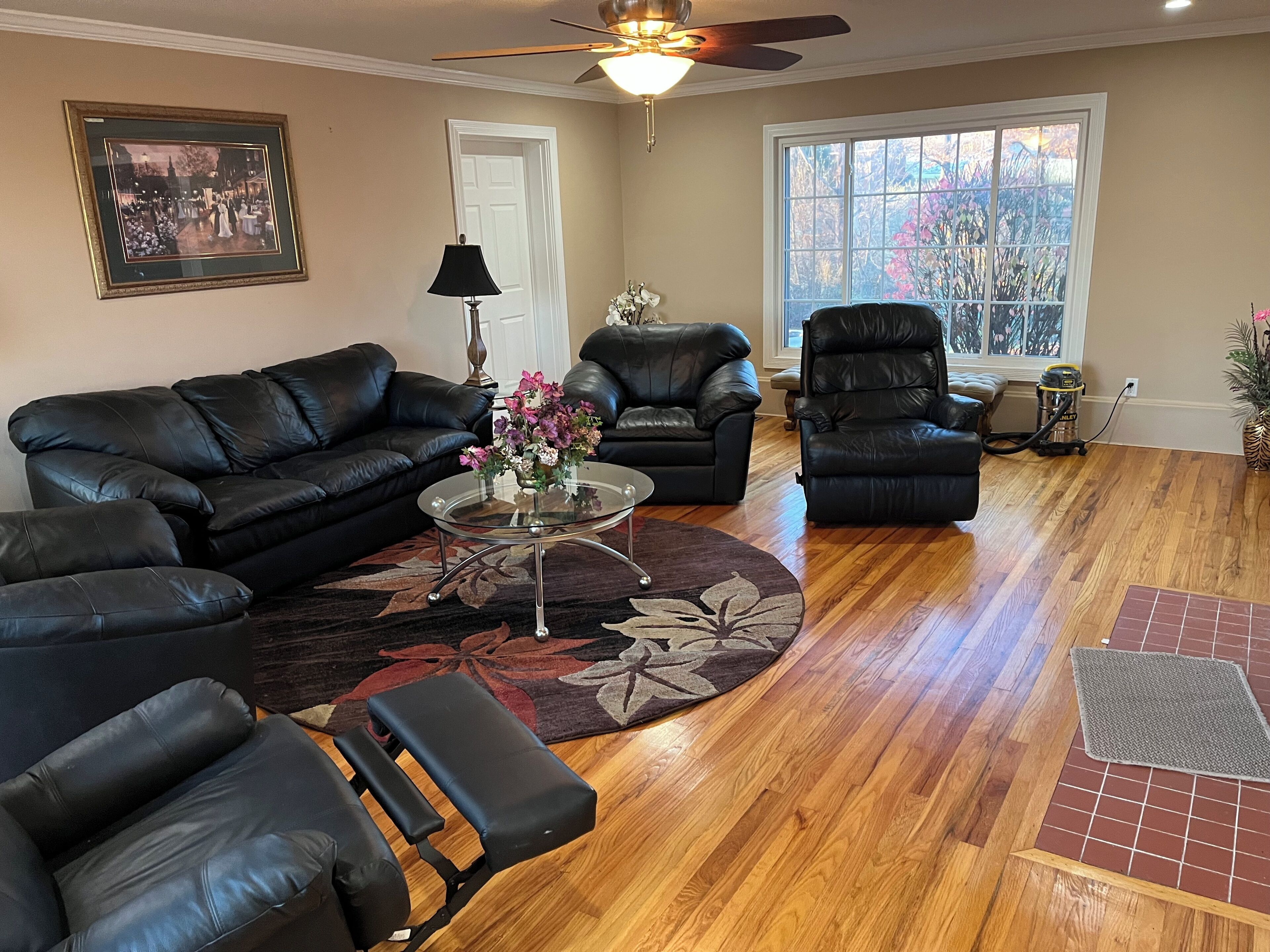 A living room with a black leather sofa, a glass coffee table with a floral centerpiece, a black leather recliner, and a large window with a view of trees.