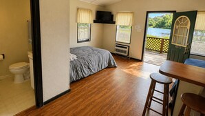 Ein Zimmer mit Bett, Toilette, einem kleinen Tisch mit Hocker, Deckenventilator und Fenster mit Blick ins Grüne.