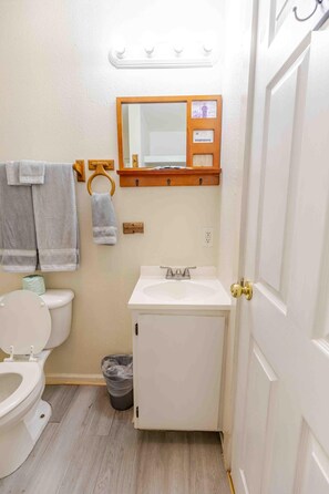 A small bathroom with a toilet, sink, and a mirror above the sink. There are towels hanging on a rack and a trash can.