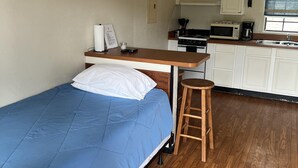 Ein kleines Zimmer mit einem Einzelbett, einem kleinen Tisch, einem Hocker, einer Küchenzeile und einem Fenster.
