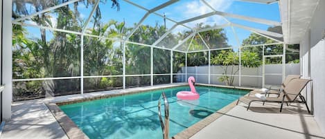 A covered outdoor swimming pool with a pink flamingo float, lounge chairs, and a view of tropical vegetation.