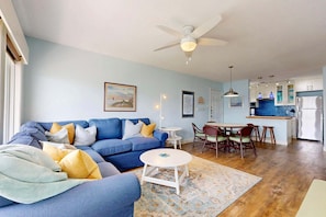 A living room with a blue sofa, a small round table, and a ceiling fan. - Shared Heated Pool, Boardwalk to Beach, Golf Cart Access (Port Aransas)