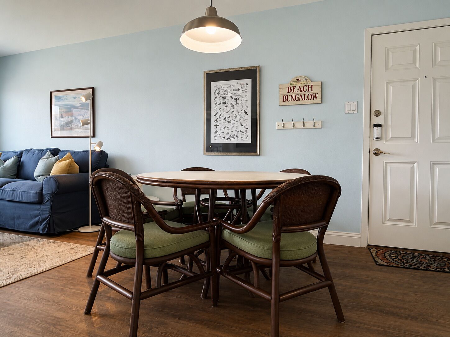 A dining area with a round table and wicker chairs, a framed wildlife illustration on the wall, a blue couch, and a door labeled 'BEACH BUNGALOW'.