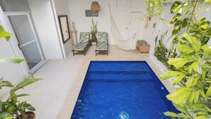 En swimmingpool med to liggestole og en hængekøje, omgivet af krukkeplanter samt en hvid væg med et spejl og en flettet kurv.